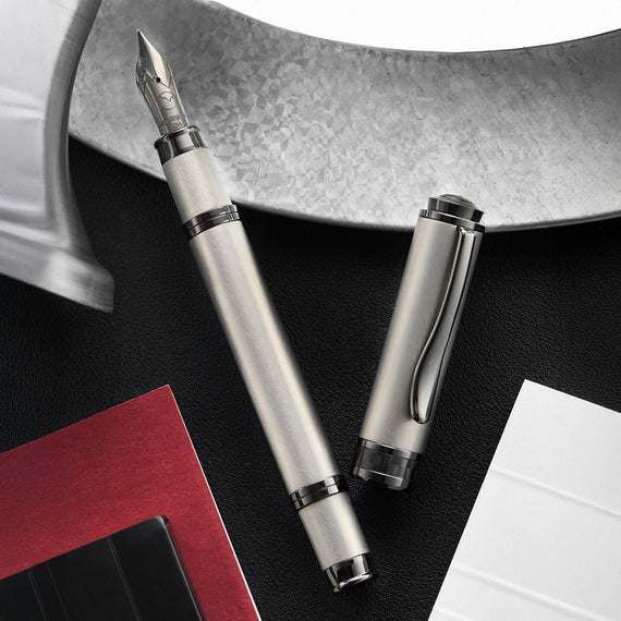 Silver fountain pen and its cap on a black surface with a book and paper in the background